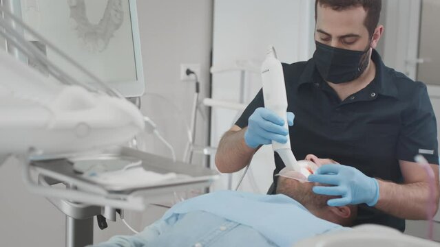 A male orthodontist doctor scans a man's teeth and jaw in the clinic's medical office. Creates a 3d model with an intraoral manual 3d scanner. Diagnosis of caries. Dentist's office in the clinic.