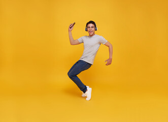 Young handsome Asian man smiling holding smartphone and jumping wearing wireless headphone listening to music isolated over yellow background.