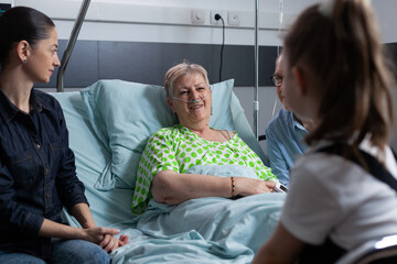 Smiling elderly woman under medical observation in hospital, chatting happily with girl. Relatives visiting senior lady during hospital stay. Happy older female patient with several people at - obrazy, fototapety, plakaty