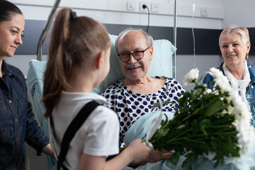 Sick grandfather receiving flowers from granddaughter in hospital room. Bedridden old man being visited by relatives in geriatric clinic room. Little girl giving flowers to bedridden elderly man.