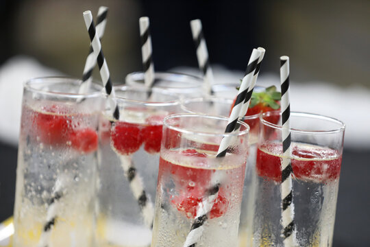 A tray of tall glass tumbler glasses filled with icy cold and clear sparkling water or soda garnished with strawberries and black and white environmentally friendly paper straws