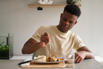 Young african man eating fried chicken, rice and plantain with tomato sauce plate in the living...