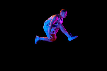 Active athletic male basketball player jumping with basketball ball isolated over dark background in purple neon light. Concept of energy, professional sport, hobby.