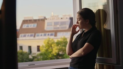 Young woman stands by the window having a sad or tough moment crying and pensive. Remembering hard times or thinking about something sad. Emotional breakdown or break up. Dramatic dark scene in house