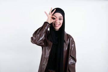 young beautiful muslim woman wearing hijab and leather jacket over white background with happy face smiling doing ok sign with hand on eye looking through finger.