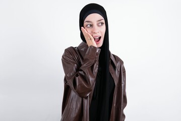 young beautiful muslim woman wearing hijab and leather jacket over white background excited looking to the side hand on face. Advertisement and amazement concept.