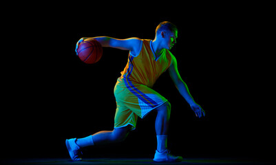 Studio shot of young active athlete, male basketball player in sports uniform in motion and action with ball isolated over dark background in neon light