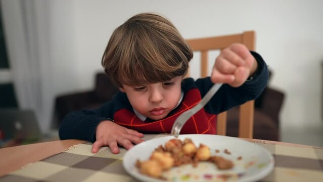 Happy small child eats food by himself with fork. One little boy eating dinner wearing sweater during winter season