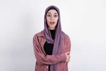 Shocked embarrassed young beautiful muslim woman wearing hijab and pink jacket over white background keeps mouth widely opened. Hears unbelievable novelty stares in stupor