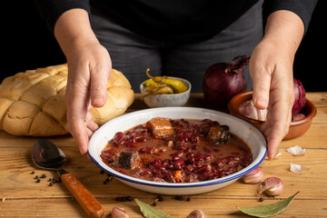 View of woman's hands with a plate of Basque beans on a rustic table with onions, garlic, bread and green chilli, selective focus, black background, horizontal