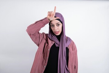 young beautiful muslim woman wearing hijab and pink jacket over white background gestures with finger on forehead makes loser gesture makes fun of people shows tongue