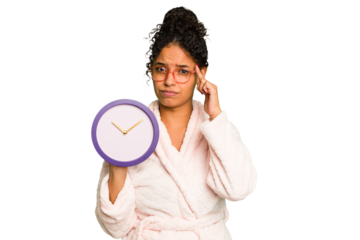 Young brazilian woman wearing a pajama holding a clock isolated pointing temple with finger, thinking, focused on a task.