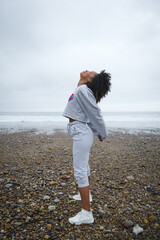 Fitness woman working out and stretching at the beach towards under autumn rainy weather. Afro hairstyle sporty black female doing breathing exercise outdoor. - obrazy, fototapety, plakaty
