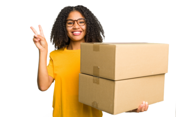 Young african american woman moving to other house while picking up a box full of things isolated joyful and carefree showing a peace symbol with fingers.