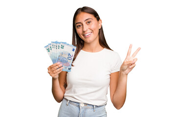 Young caucasian woman holding a banknotes isolated joyful and carefree showing a peace symbol with...