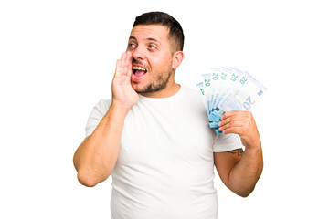 Young caucasian man holding a banknotes isolated shouting and holding palm near opened mouth.