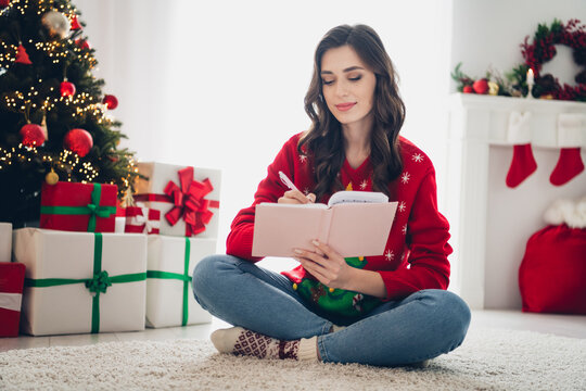 Photo of young curly hair woman sit floor hold copybook writing wish list her ideas for new year gifts near xmas tree atmosphere decoration indoors