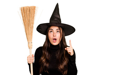 Young caucasian woman dressed as a witch holding a broom isolated on green chroma background pointing upside with opened mouth.