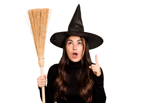 Young caucasian woman dressed as a witch holding a broom isolated on green chroma background pointing upside with opened mouth.