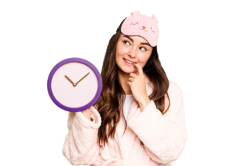 Young caucasian woman holding a clock wearing a pajama isolated on green chroma background relaxed thinking about something looking at a copy space.