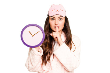 Young caucasian woman holding a clock wearing a pajama isolated on green chroma background keeping a secret or asking for silence.