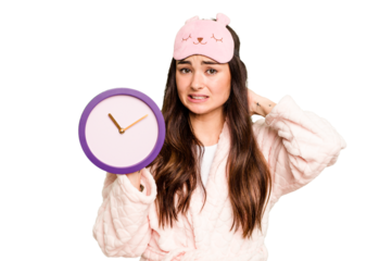 Young caucasian woman holding a clock wearing a pajama isolated on green chroma background touching back of head, thinking and making a choice.