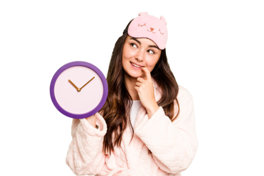 Young caucasian woman holding a clock wearing a pajama isolated on green chroma background relaxed thinking about something looking at a copy space.