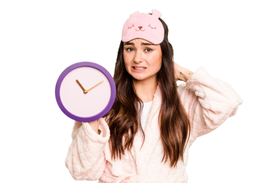 Young caucasian woman holding a clock wearing a pajama isolated on green chroma background touching back of head, thinking and making a choice.