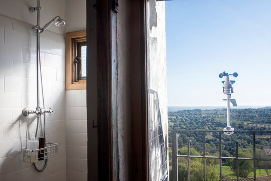 Lavabo con ducha de dise&ntilde;o en la mitad de la fotograf&iacute;a y balc&oacute;n con vistas en la mitad derecha con aparato metereol&oacute;gico, d&iacute;a soleado. Casa rural de pueblo con vistas a campos  y bosque.