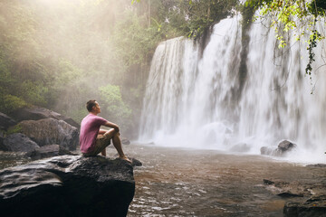 Man sitting on rock in front of high waterfall in mountains in tropical landscape in Camobodia..