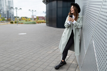 a stylish charismatic girl in an autumn coat and a black hat walks around the city next to modern...