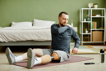 Full length portrait of man with prosthetic leg doing stretching exercises on yoga mat at home