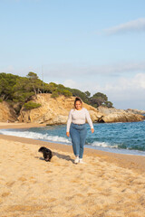 Mujer joven jugando en la playa con su bonito perro negro llamado Loki. Corriendo, sonriendo, queriéndose y abrazándose en actitud feliz y disfrutando de un agradable día - obrazy, fototapety, plakaty