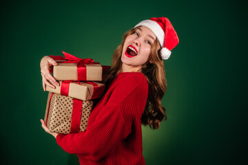 Happy young woman in santa hat holding stack of gift boxes isolated on green background. Christmas shopping concept