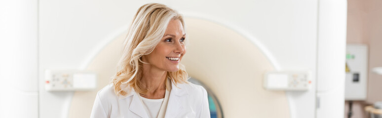 happy blonde radiologist looking away near computed tomography machine, banner.