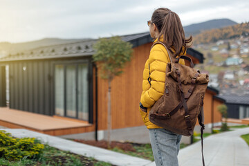 Young female in yellow down jacket with leather retro tourism backpack have a walk in nature background near eco houses