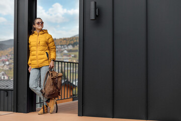 Young female in yellow down jacket with leather retro tourism backpack have a walk in nature background near eco houses