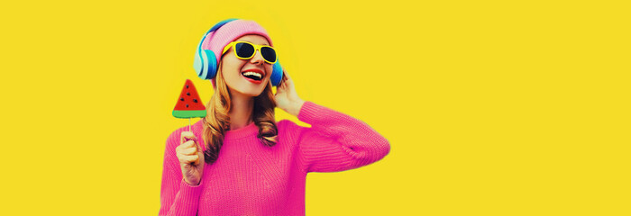 Summer fresh colorful portrait of happy laughing young woman in headphones listening to music with fruit juicy lollipop or ice cream shaped slice of watermelon on yellow background