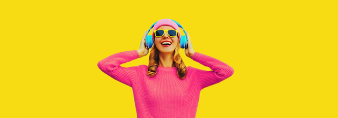 Portrait of happy smiling young woman with headphones listening to music wearing colorful pink sweater on yellow background