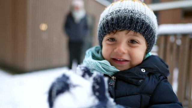One small boy standing outside during winter season playing with snow. Portrait of a happy child wearing beanie and coat holding snowball