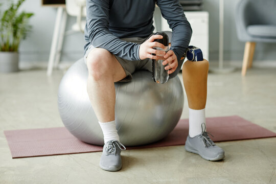 Low section closeup of man with prosthetic leg sitting on fitness ball and holding water bottle after home workout