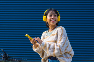 Portrait of Asian girl listening to music with yellow headphones on a blue college background