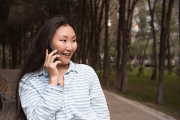 Young happy asian womanhaving a talk by phone walking in city park at summer