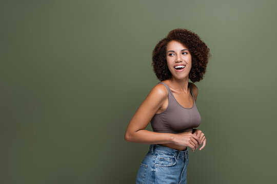 Cheerful african american model in top and jeans looking away isolated on green.