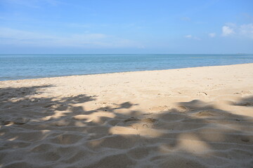Sunlit beach with tree shade on the sand. Calm sea, bright blue sky, white clouds, sea without waves, suitable for relaxation.
