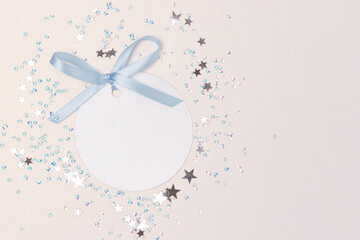 Round empty tag with blue tied ribbon bow. Stars confetti on a glittering silver background.
