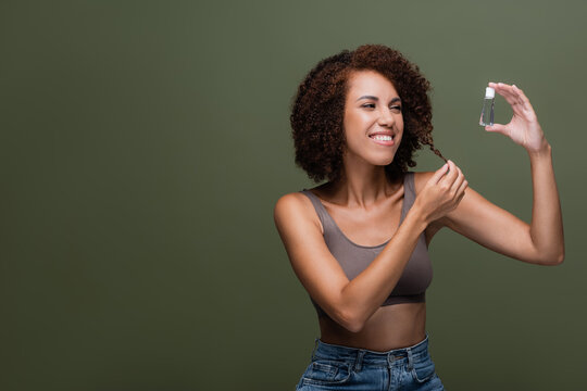 Positive african american woman holding cosmetic oil and touching curly hair isolated on green.