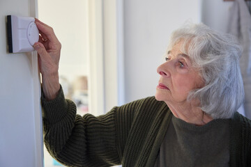 Worried Senior Woman Turning Down Central Heating Thermostat At Home In Energy Crisis