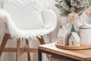 A bouquet of fir trees, a plaid in a wicker basket and Scandinavian white houses on a wooden table in the home interior of the living room. A cozy concept of festive home decoration.