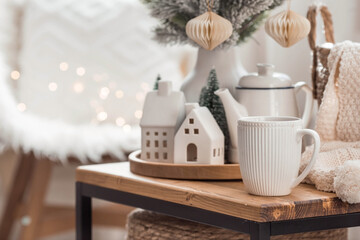 A bouquet of fir trees, a plaid in a wicker basket and Scandinavian white houses on a wooden table in the home interior of the living room. A cozy concept of festive home decoration.